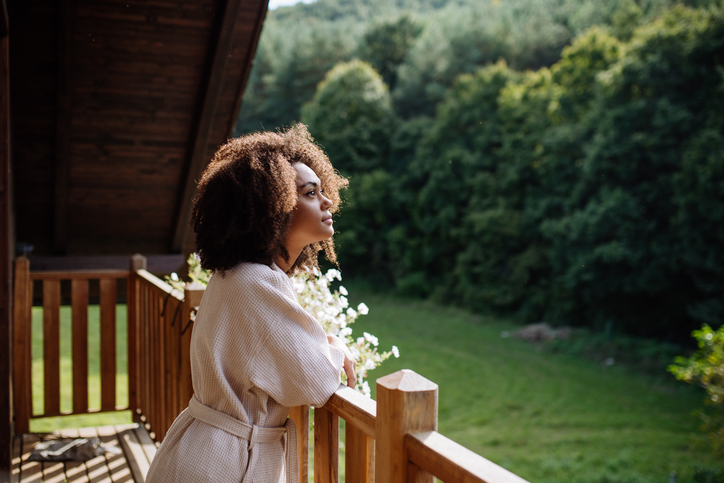 Peaceful morning for young woman standing on balcony in bathrobe, looking at beautiful nature around her, smiling.